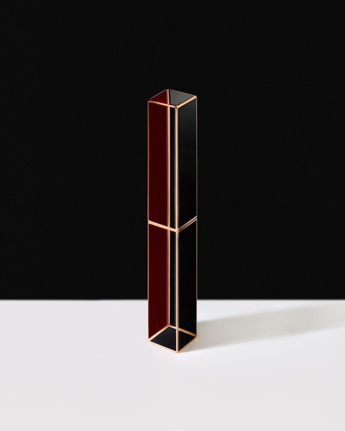 A slim rectangular lipstick tube with a geometric design, featuring alternating panels of deep burgundy red and glossy black, outlined with gold metal trim, standing on a white surface against a black background.